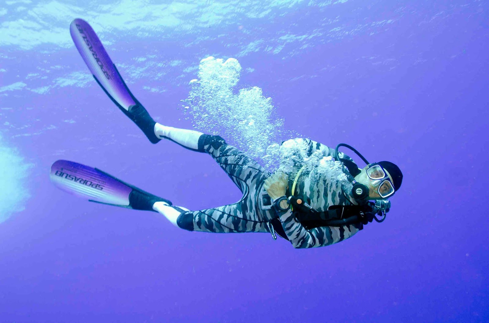 A diver in a camouflage wetsuit finning through open blue water with a trail of bubbles